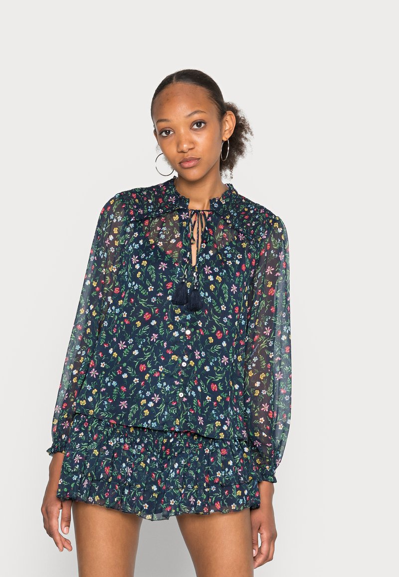 Blusa a fantasia floreale di un blu navy scuro con tessuto trasparente, orlo arricciato, maniche lunghe e fiocchetto al collo. Abbinata a una gonna arricciata coordinata.