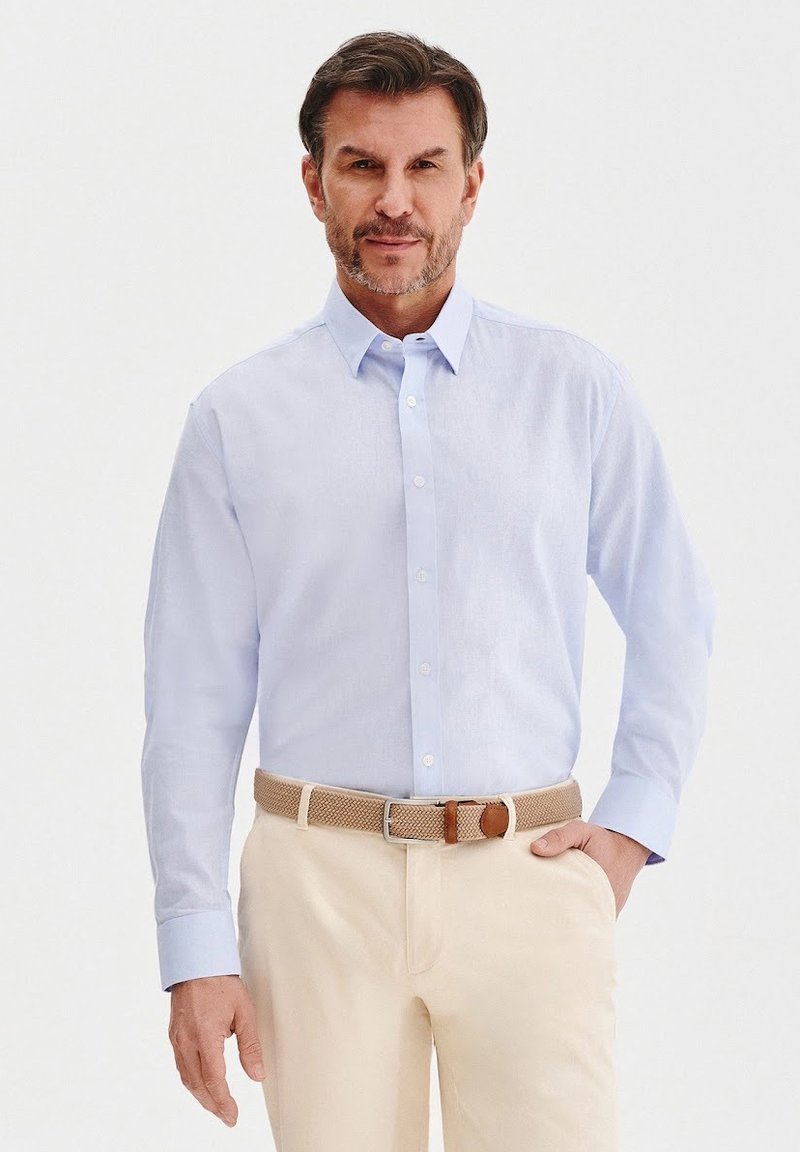 Jasnoniebieska koszula zapinana na guziki z kołnierzykiem, z długimi rękawami i białymi guzikami, połączona z kremowymi spodniami i plecionym brązowym paskiem.