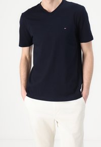 Námořnická modrá bavlněná tričko s výstřihem do V a krátkými rukávy. Malé logo s červenými a bílými akcenty umístěné na levé hrudi.