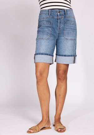 Frau trägt hochgekrempelte blaue Jeansshorts mit großen Fronttaschen, gestreiftes Oberteil und braune offene flache Sandalen vor einfarbigem Hintergrund.