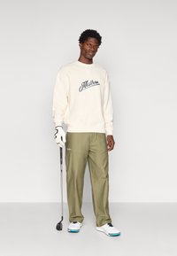 Hanorac crem cu logo "Malbon", pantaloni verzi măslinii, încălțăminte albă și un club de golf. Ansamblul este casual, dar orientat spre sport.