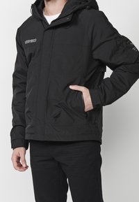 Svart vadderad jacka med huva, utrustad med blixtlåsfickor på sidorna, elastiska ärmmuddar och logodetaljer på bröstet och ärmen. Slät tygkvalitet.