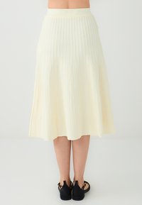 Jupe plissée jaune clair avec une taille élastique, atteignant mi-mollet, présentant une texture lisse et une silhouette fluide. Sandales noires visibles.