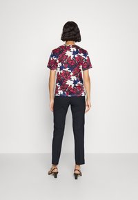 Květinová košile s krátkým rukávem s jasně červenými a fialovými květinami na bílém pozadí, v kombinaci s úzkými tmavými kalhotami a sandály na blokovém podpatku.