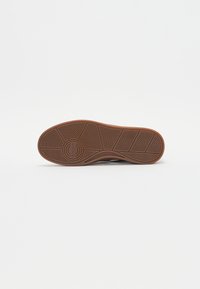 Brown rubber shoe sole featuring a zigzag tread pattern and a circular logo imprinted in the center. Smooth surface with rounded edges.