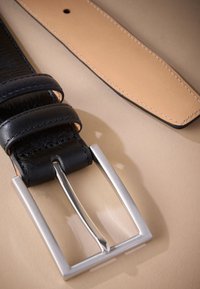 Ceinture en cuir noir avec boucle en métal argenté ; elle possède deux passants et un dessous lisse couleur beige. Des surpiqûres longent les bords en guise d'accent.