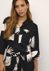 Chemise noire à boutons avec imprimé de feuilles de palmier blanches, dotée d'une poche sur la poitrine et d'une taille nouée. Tissu lisse, manches 3/4, coupe décontractée.