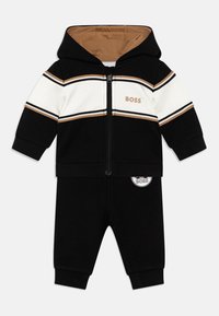 Ensemble de survêtement à capuche avec des blocs de couleurs noir et blanc et des rayures brunes. Comprend une fermeture éclair à l'avant, un détail de logo et un pantalon noir assorti.