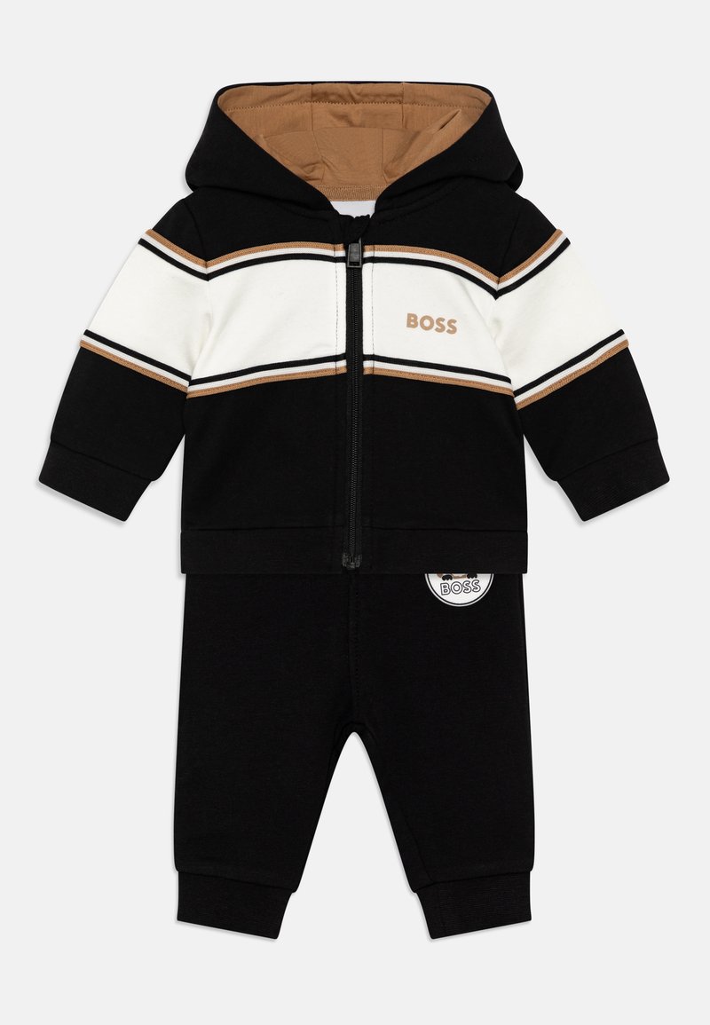 Ensemble de survêtement à capuche avec des blocs de couleurs noir et blanc et des rayures brunes. Comprend une fermeture éclair à l'avant, un détail de logo et un pantalon noir assorti.