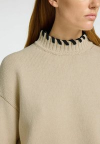 Beige pletený svetr s vysokým ribbed límcem a černými akcenty. Měkká textura a volný střih, ideální pro vrstvení.