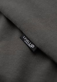 Gros plan sur un tissu gris foncé avec une étiquette de vêtement noire cousue dans la couture, portant l'inscription "LEVEL UP ombre".