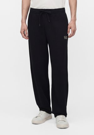 Homme portant un pantalon de survêtement noir avec cordon et des baskets blanches, debout sur un fond blanc uni.