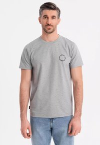 Camiseta de algodón gris con pequeñas motas negras, escote redondo, mangas cortas y un logo circular en la parte izquierda del pecho.