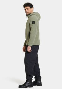 Olivgrön fleecejacka med huva, med en svart logotyp-patch, sidofickor med dragkedja, och bärs med svarta cargobyxor och kängor.