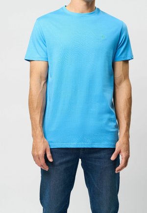 Mężczyzna w gładkim, jasnoniebieskim t-shircie z krótkim rękawem i ciemnoniebieskich dżinsach, stojący na jasnym tle.