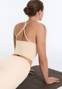 Femme en tenue de sport beige avec des bretelles tressées effectuant la posture de yoga du chien tête en haut sur un tapis d'exercice sombre, vue de dos.