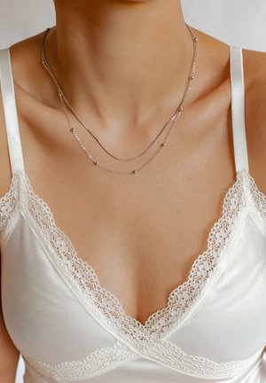 Colliers superposés en argent avec des chaînes délicates et texturées, ainsi que de petites perles rondes, portés sur un fond de tissu blanc avec des dentelles.