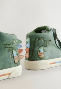 Zöld bőr magas szárú cipők krokodil mintával, texturált díszítésekkel, színes szövet pikkelyek a sarokrészen, fehér gumitalppal csíkokkal.