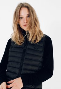 Sort quiltet vest med lynlås foran, vandret quiltning og rundt logo. Båret over en langærmet sort top med en glat stofstruktur.