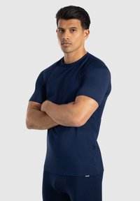 Granatowa, krótka koszulka sportowa z gładkiego materiału z okrągłym dekoltem i logo na dolnym hemie. Dopasowany fason.