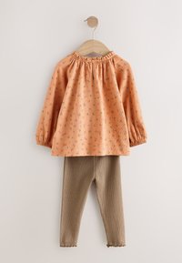 Haut à motifs floraux pêche avec manches bouffantes, associé à des leggings côtelés beige, présenté sur un cintre en bois sur un fond neutre.
