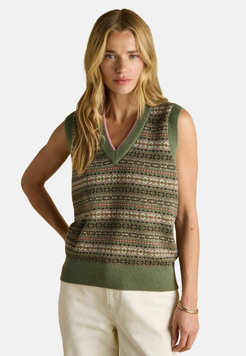 Pikkade blondide juustega naine, kes kannab roheline mustriga säärest vabalt langevat kampsunit ja kreemikaid pükse, seisab ühe käega puusa peal.