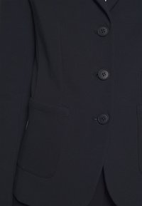Blazer navy con cuatro botones negros, solapas de muesca y dos bolsillos frontales con una textura suave y ajustada.