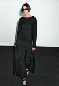 Maglia nera a maniche lunghe in maglia abbinata a una gonna nera plissettata e fluida. La modella indossa occhiali da sole neri e cinturini alla caviglia. Tessuti lisci e testurizzati.