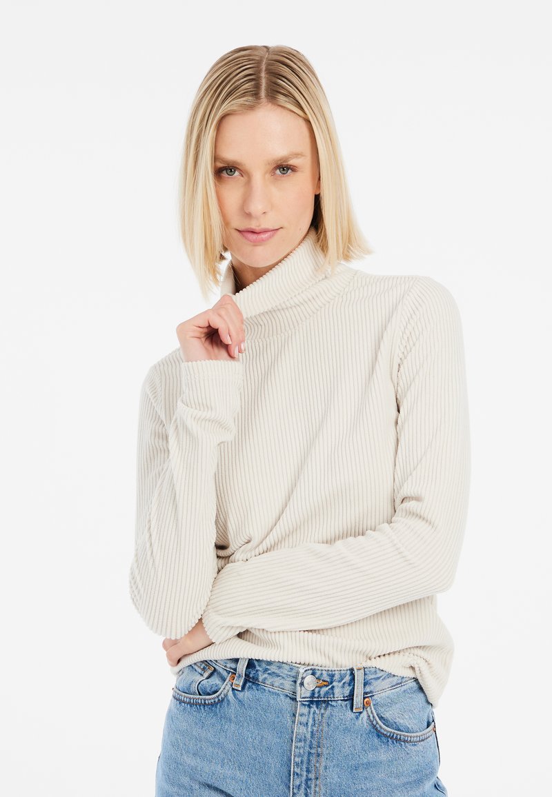 Pull à col roulé beige à côtes avec des poignets ajustés, coupe décontractée et texture douce, associé à un jean en denim bleu taille haute.