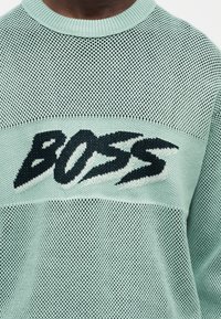 Άτομο που φοράει ανοιχτό πράσινο πλεκτό πουλόβερ με μεγάλο μαύρο και άσπρο κείμενο "BOSS" στο στήθος σε έντονα, στιλιζαρισμένα γράμματα.