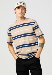 Camiseta beige a rayas con rayas horizontales en azul marino y blanco, diseño de cuello redondo, combinada con una gorra clara que presenta texto en azul marino.