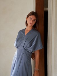 Siva, plisirana midi haljina s V-izrezom, kratkim kimono rukavima i teksturiranim materijalom. Naglašena prilagođenim strukom i lepršavom suknjom.
