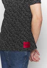 Sort kortærmet skjorte med hvid geometrisk mønster, med en rød Hugo-label ved kanten. Bæres sammen med blå denim jeans.