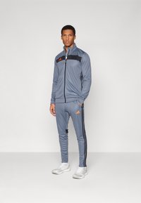 Heren sporttracksuit in grijs, met zwarte accenten en oranje logo. Jack met rits, aansluitend ontwerp en contrasterende zijstrepen.