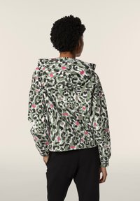 Giacca verde e nera a fantasia con dettagli rosa, cappuccio, polsini elastici e vestibilità rilassata. Il tessuto ha una texture liscia e un design leggero.