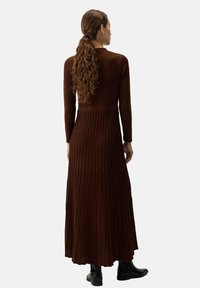 Naine pika lokkis juustega, pruunis ribakujulises pika varrukaga maksikleidis ja mustades pahkluu saapades, seisab seljaga näkku tasase taustaga.