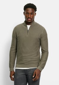 Olivgrüner Halbzip-Pullover mit strukturiertem Rippdesign, langen Ärmeln und einem enganliegenden Bund. Über einem weißen Hemd getragen.