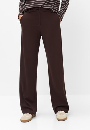 Personne portant un pantalon large marron foncé avec les mains dans les poches et des baskets beige sur fond blanc.