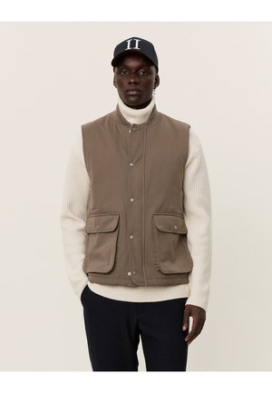 Gilet marrone senza maniche con due tasche frontali, realizzato in tessuto morbido. Indossato sopra un maglione bianco a coste. Berretto blu marino con scritte bianche.