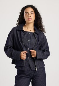 Donkerblauwe gestreepte denimjack met een ritssluiting, lange mouwen en een kraag. Voorzien van grote zilveren knopen en een cropped pasvorm.