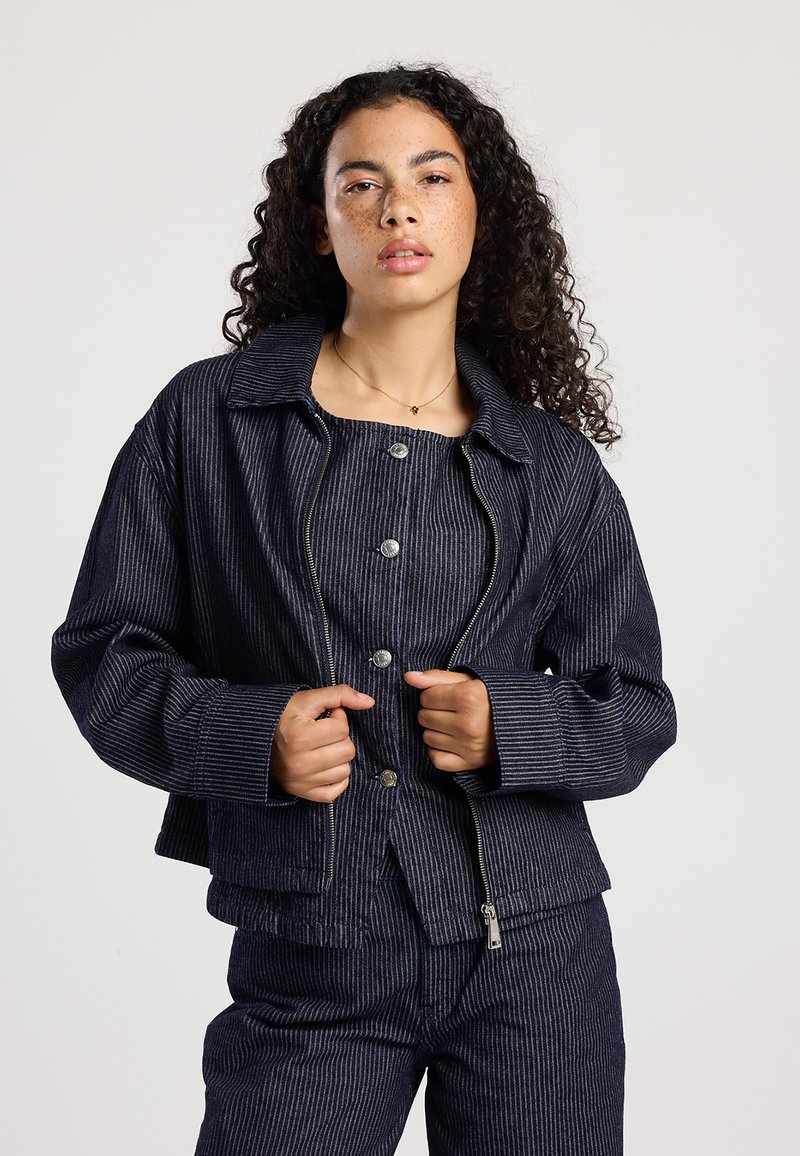Donkerblauwe gestreepte denimjack met een ritssluiting, lange mouwen en een kraag. Voorzien van grote zilveren knopen en een cropped pasvorm.