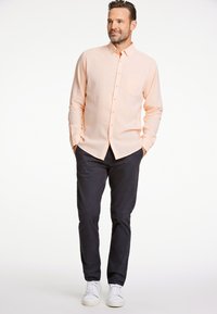 Camisa de botón color melocotón claro con un bolsillo en el pecho, combinada con pantalones azul marino oscuros y zapatillas blancas. Tela suave con una textura sutil.
