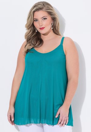 Donna con lunghi capelli biondi ondulati, che indossa un top verde acqua senza maniche e orecchini a goccia di perle, sorridente su sfondo bianco.