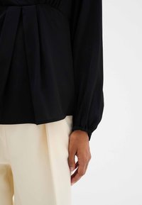 La blusa negra presenta mangas largas y fruncidas, con un acento plisado en la cintura, combinada con pantalones crema de corte ajustado.