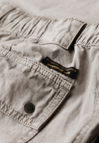 Helle graue Stoffshorts mit einem elastischen Bund. Verfügt über eine Klapptasche mit einem schwarzen Etikett und einem silbernen Druckknopf-Detail. Glatte Textur.