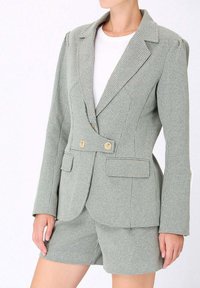 Blazer à carreaux verts et blancs avec deux boutons dorés, deux poches avant et une coupe ajustée, associé à un short assorti. Texture de tissu lisse.