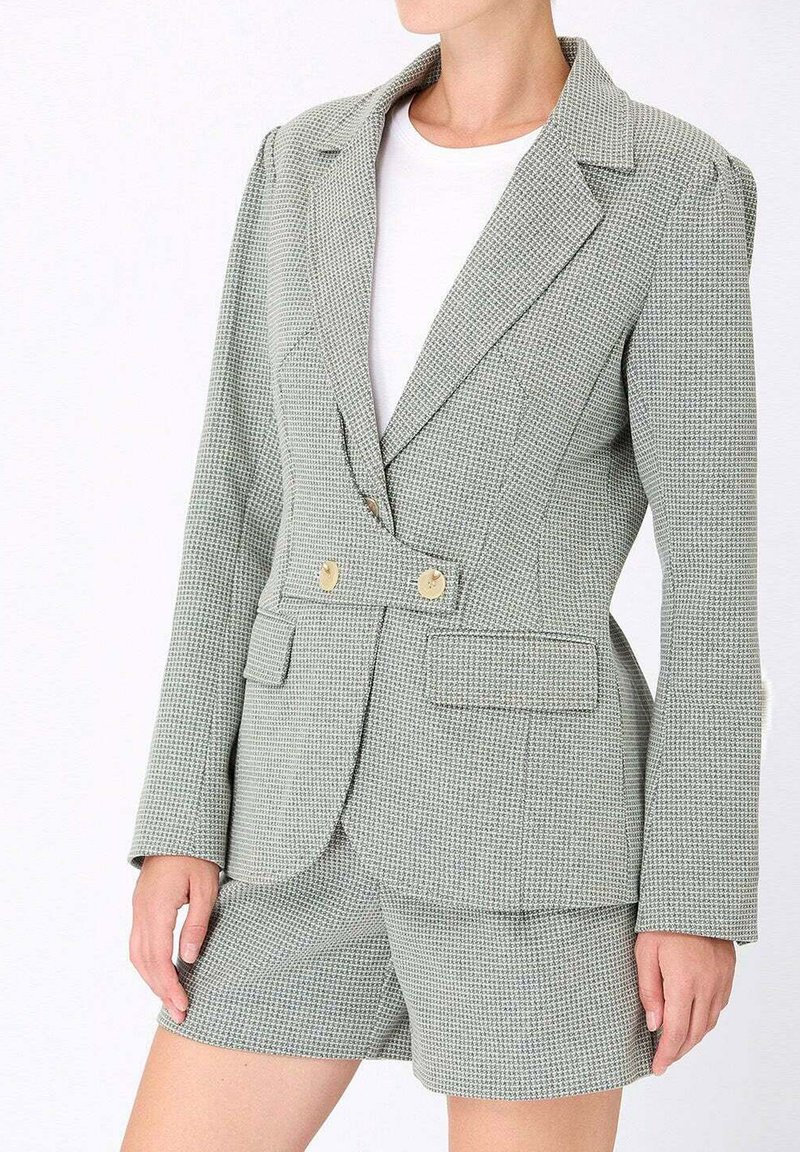 Blazer à carreaux verts et blancs avec deux boutons dorés, deux poches avant et une coupe ajustée, associé à un short assorti. Texture de tissu lisse.