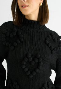 Black knitted sweater featuring a high neck, textured heart patterns made of pom-poms, and ribbed detailing on the sleeves.