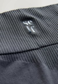 Tessuto grigio a costine con un pannello liscio che presenta un logo del marchio riflettente. Il materiale ha una texture morbida con motivi di lavorazione a maglia variabili.