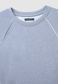 Sweatshirt gris clair avec un col rond, des manches raglan, une texture subtile et des détails de couture blancs contrastants. Étiquette de la marque visible.